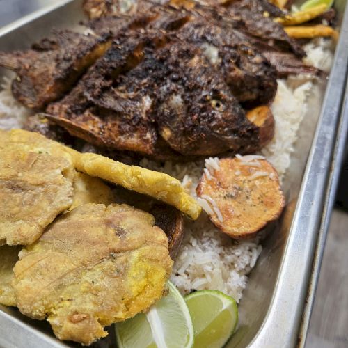 A tray of smoked meats and fried sides over rice, with slices of lime wedges and some grilled onions on the side.