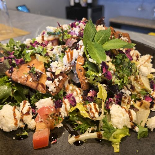 A colorful tossed salad with greens, feta, tomatoes, chicken or tofu, herbs, and a drizzle of balsamic glaze on a dark plate.