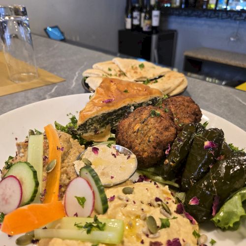 A plated meal with hummus, grilled vegetables, cucumber, radish, carrot, herbs, flatbread, and falafel or croquettes on a chic restaurant bar. Top it at 140 characters, always ending the sentence.