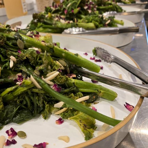 Assorted sautéed greens (kale or spinach) with asparagus, almonds, and pink flower petals on large white plates, at a buffet-style setup.