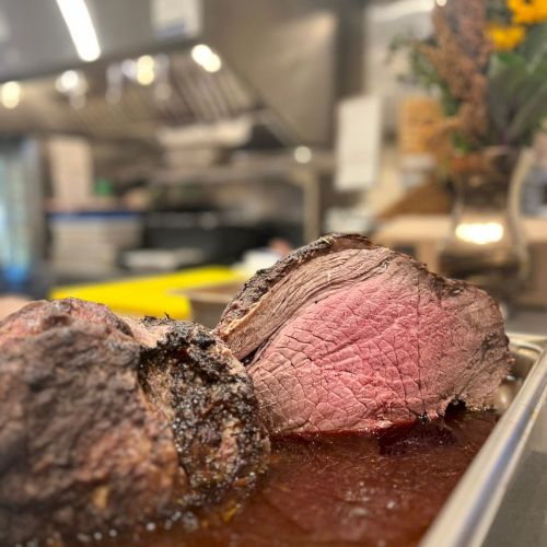 A close-up of perfectly seared roast beef slices, rare to medium-rare, resting in their juices on a tray in a kitchen.