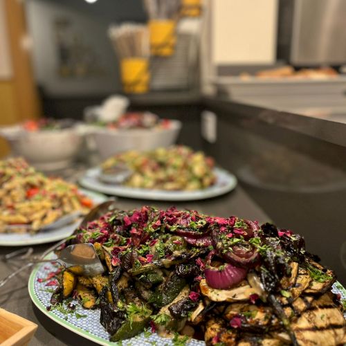 A plate of grilled vegetables and peppers with char marks, greens and red onions, in a buffet line with other dishes in the background.