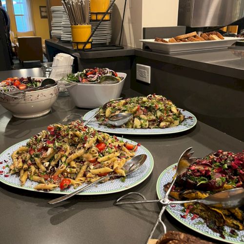 A buffet spread with sliced roasted meat, noodles, roasted vegetables, fresh salads, bowls of toppings, bread, and utensils on a counter.
