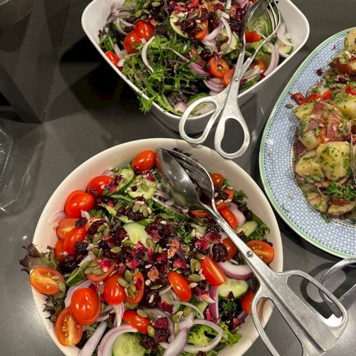 Two fresh salads with mixed greens, cherry tomatoes, cucumbers, red onions, and seeds in bowls; a third plate with chopped veggies/peppers nearby.