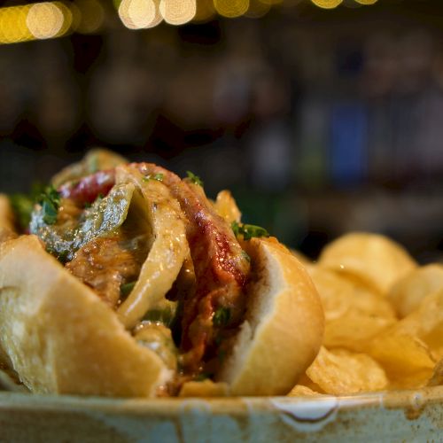 A loaded hot dog nestled in a soft bun with peppers, onions, and greens, plus a side of crispy potato chips on a plate.