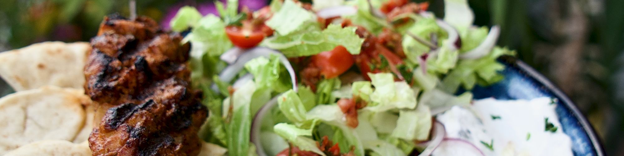 A colorful plate with grilled skewers, crispy fried onion rings, lettuce salad, tortilla chips, and a drizzle of sauce in a garden setting.