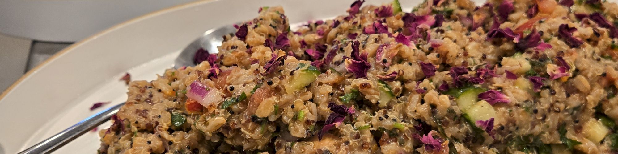 A spread of quinoa or bulgur crunch topped with herbs and pink rose petals, arranged in large oval plates, likely a communal dish, vibrant and colorful.