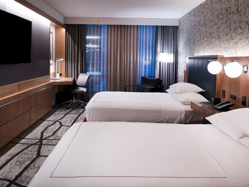 A modern hotel room with two neatly made beds, bedside lights, a wall TV, and large curtains by the window.