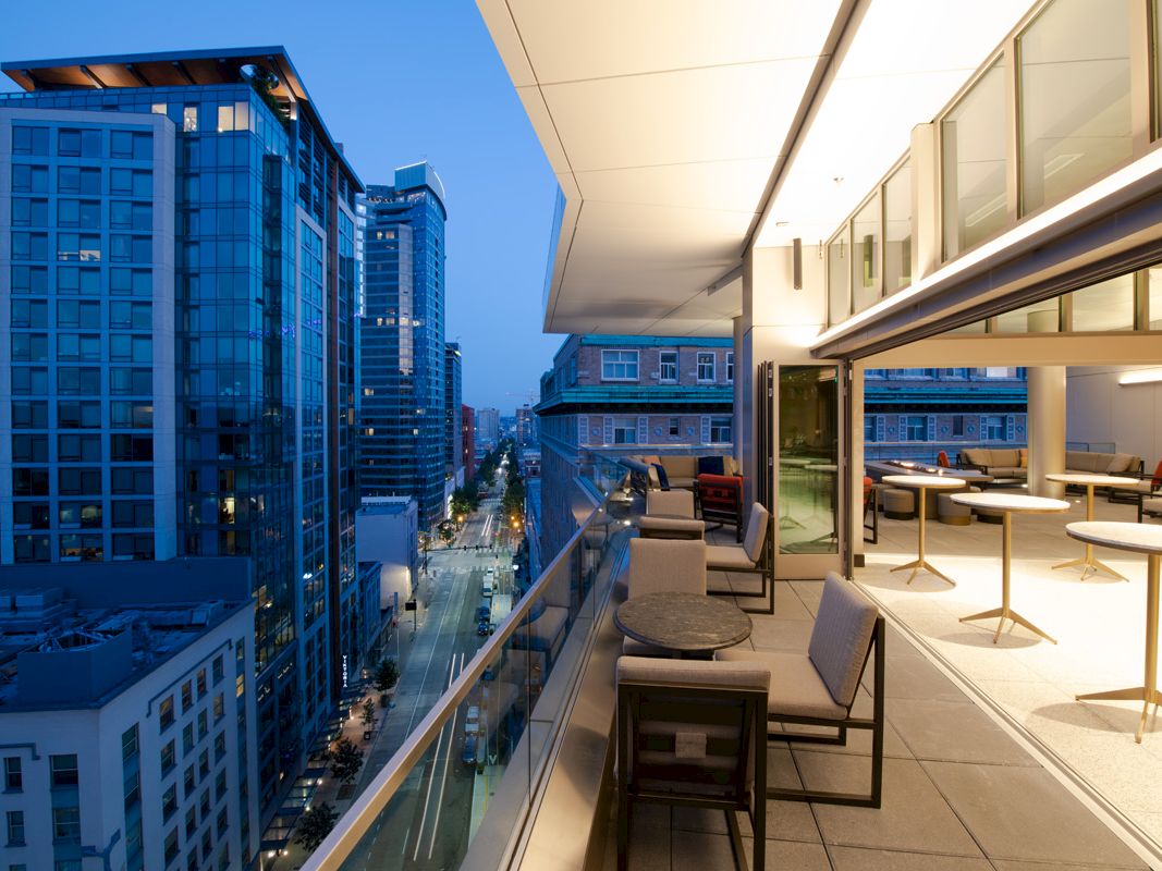 A modern high-rise rooftop terrace with lounge seating, overlooking a city street lined with tall buildings under blue evening sky.